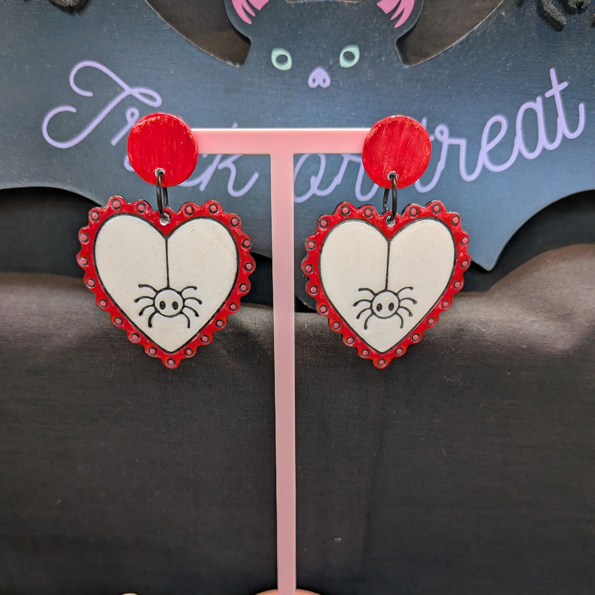 Heart-shaped dangle earrings with red borders, white centers, spider illustrations, and red circular studs, displayed on a pink stand against a Halloween-themed backdrop.